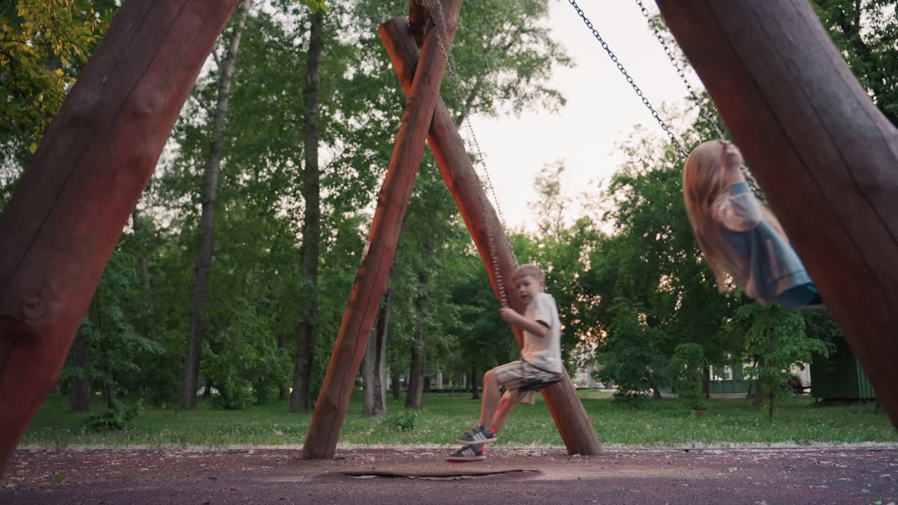 side view of siblings and friends swinging opposite directions in leafy park at dusk, laughter and carefree motion under soft sunset light, capturing playground fun in serene summer evening