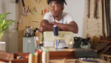 nachdenklicher afroamerikanischer handwerker mit dreadlocks, der sich auf eine nähmaschine in einer lederwerkstatt stützt