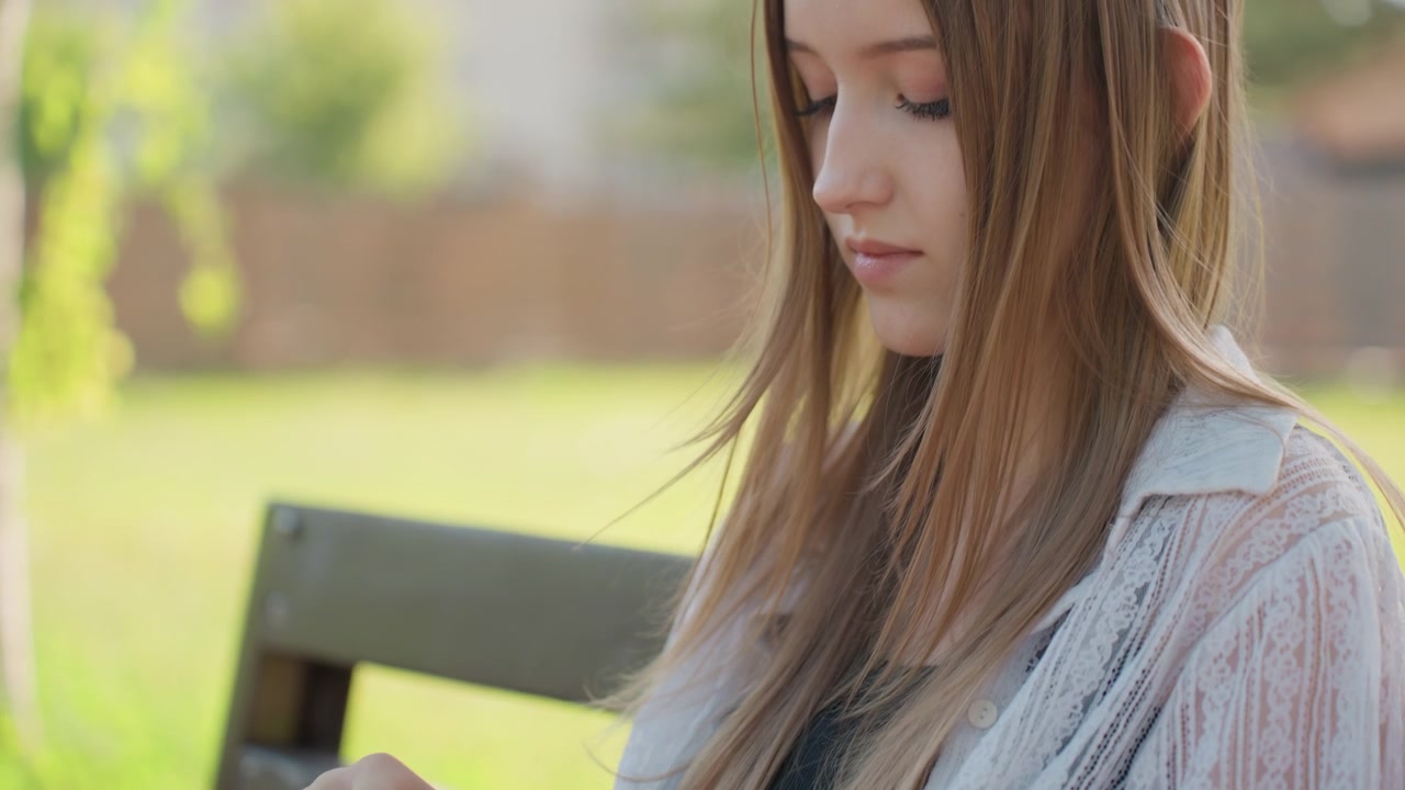 Close-up de jovem sentada ao ar livre em um banco, envolvida em crochê com fio rosa. A mulher veste tênis e meias brancas. O foco permanece firmemente em suas mãos trabalhando o tecido enquanto sentada.