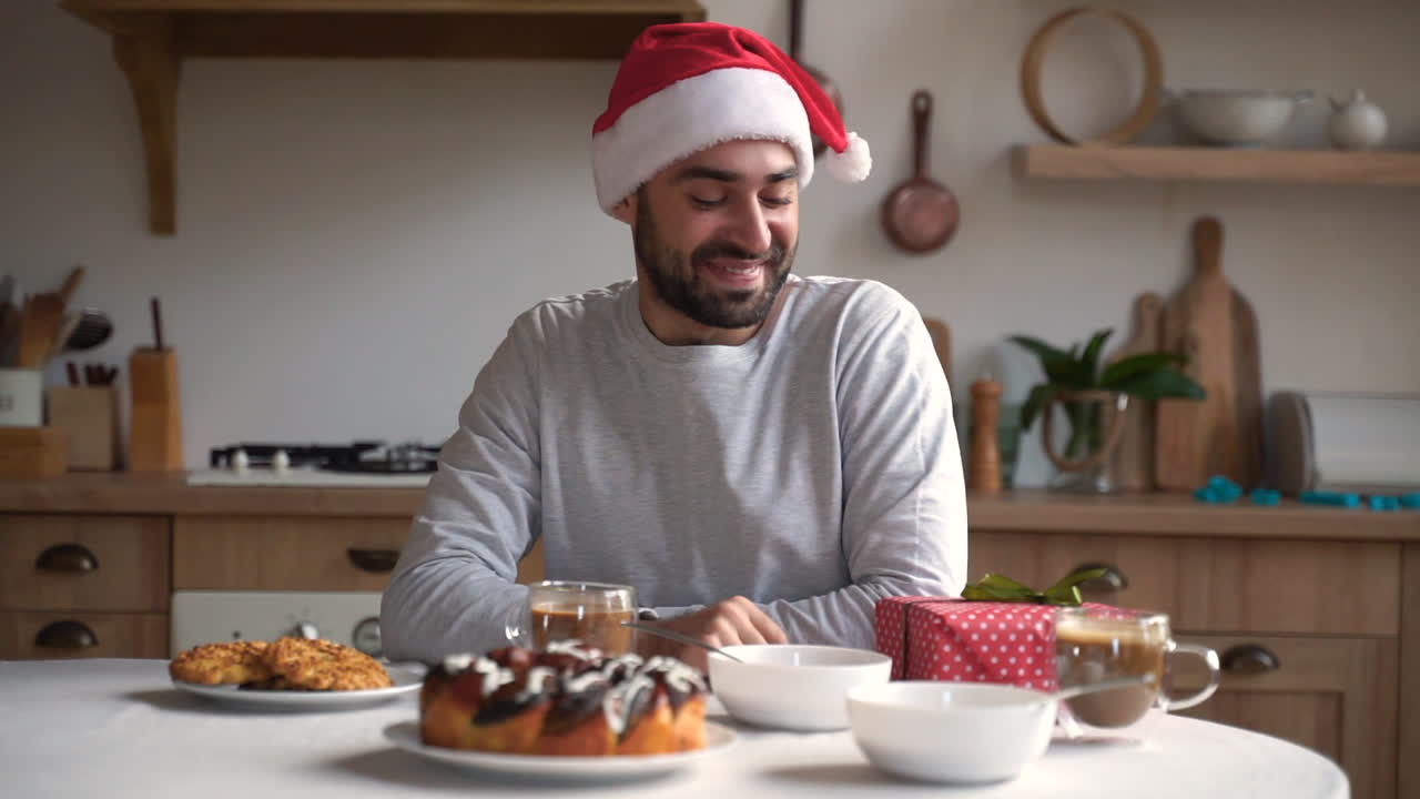 hombre emocionado sostiene un regalo de navidad mientras desayuna con sombrero rojo de santa
