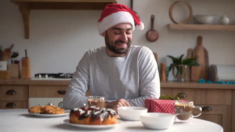 Excited-Man-Holds-Christmas-Present-While-Having-Breakfast-Wearing-Red-Santa-Hat