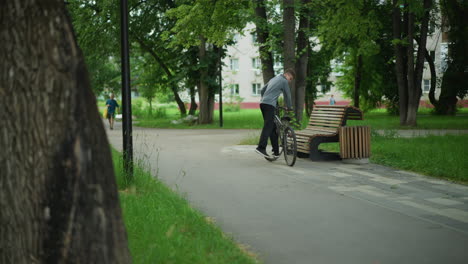 young boy in gray top rides bicycle through peaceful, tree-lined park, stops by wooden bench to sit down, another boy with yellow bag passes by, surrounded by lush greenery and residential building