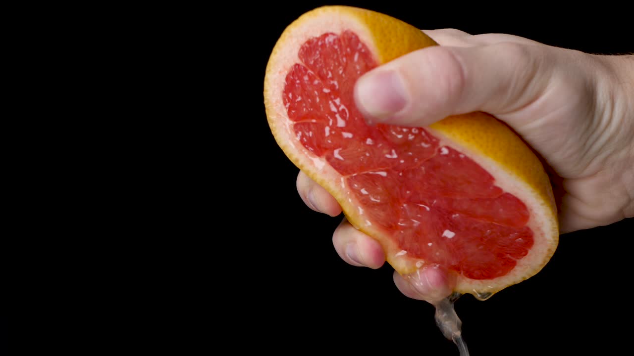 cámara lenta exprimiendo un pomelo medio cortado con una mano blanca con mucha presión sobre un fondo negro