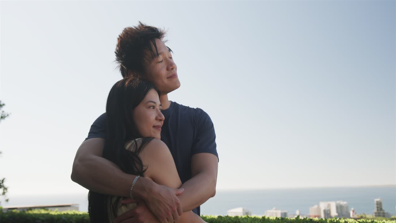 Diverse couple embracing on rooftop terrace by trimmed hedge and railing, skyline and ocean visible