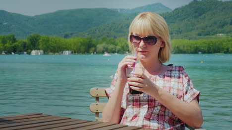 young tourist woman relaxing in a beautiful location by the lake and mountains drinks coke from a gl