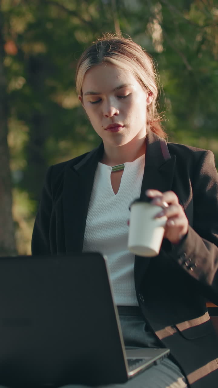 Una remote worker concentrata prende una bevanda e la sorseggia mentre lavora al laptop all'aperto, con la luce del sole che illumina il suo viso circondato da alberi e vegetazione