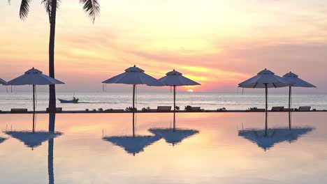 a view from across a resort swimming pool framed by beach umbrellas, a lone fishing boast travels across a stunning colorful sunset