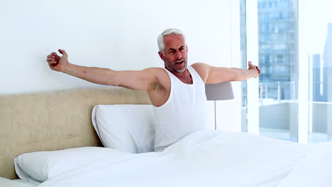 Smiling-man-yawning-and-stretching-sitting-on-bed