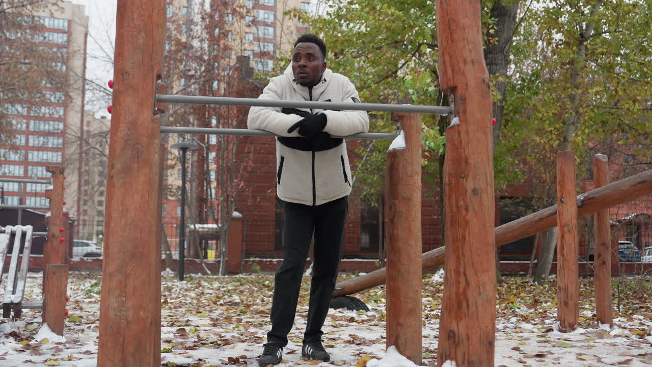 Young adult leans on iron bar looking exhausted after workout, dressed in warm fleece jacket and gloves, catching breath in cold outdoor setting with snow-covered ground