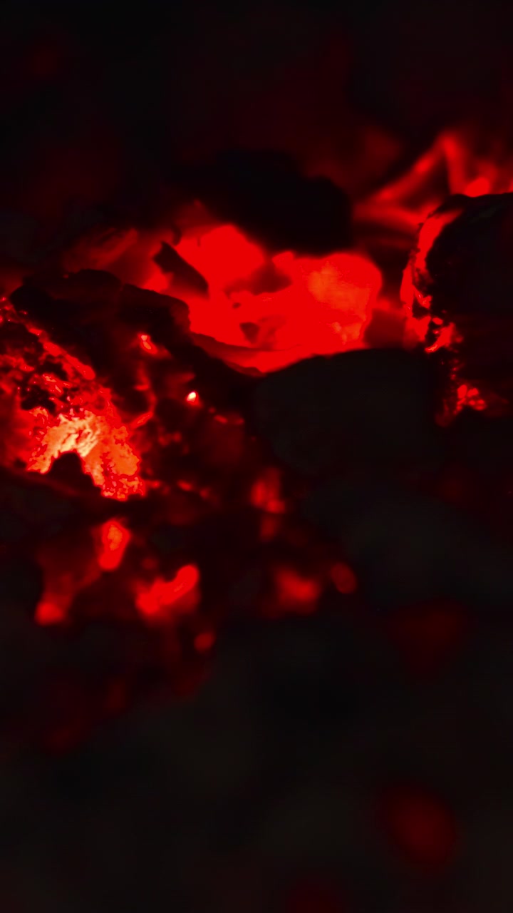 Nahaufnahme: Rote Glut in schwachem Licht, glühende Fragmente bewegen sich in rauchiger Dunkelheit mit leuchtend roten Stellen und haptisch