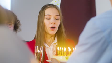 Mujer-Feliz-Soplando-Velas-En-El-Pastel-Y-Brindando-Con-Sus-Colegas-Multiétnicos-En-La-Oficina