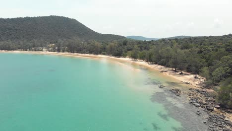 rocky golden sandy shoreline with lush green hills encircling the shallow turquoise waters of m'pai bay, koh rong sanloem, cambodia - aerial low angle fly-over shot