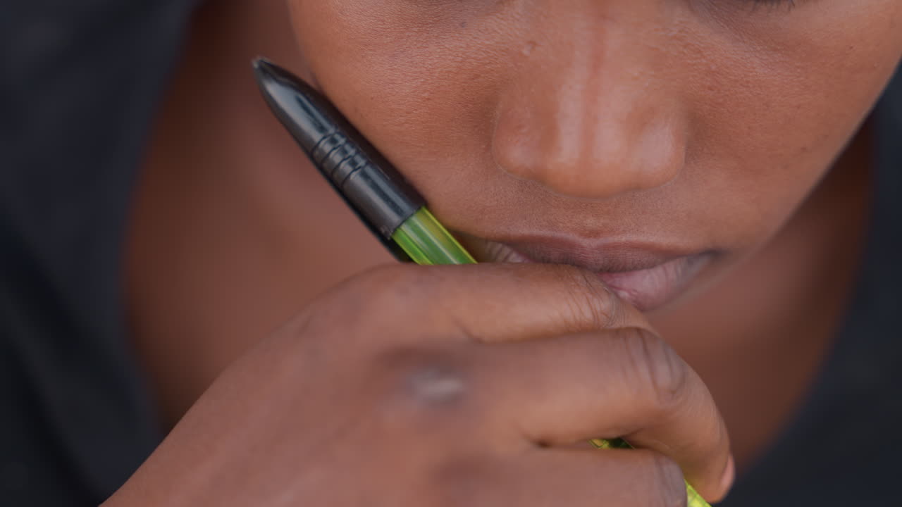 Primer plano de estudiante negro/a sosteniendo un bolígrafo en los labios, cabello trenzado, encuadre íntimo que muestra boca pensativa y agarre con la yema del dedo, detalle de bolígrafo con tinta, introspección tranquila y un estado de ánimo de decisión incierta.