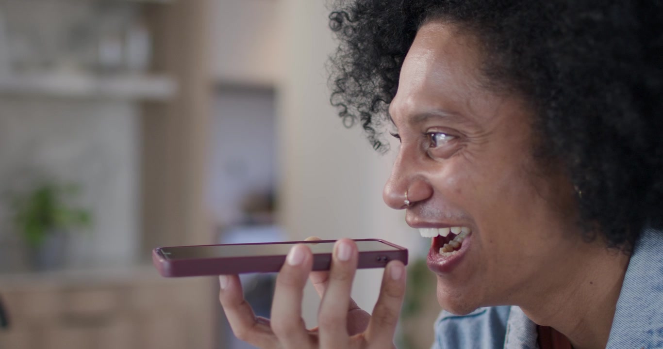 Afroamerikansk mand løfter telefonen, filmer, smiler og griner i køkkenet, iført denimskjorte, med plads til tekst.