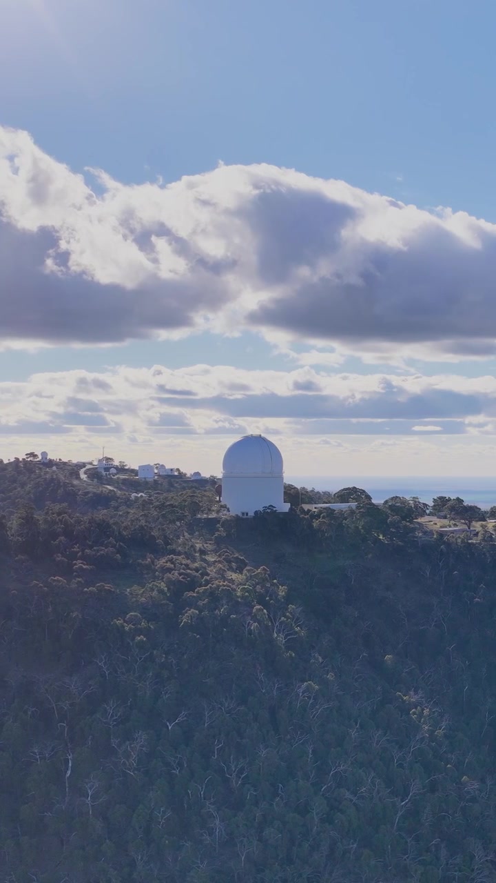 Riprese con drone che panoramicano su una cresta montuosa boscosa con una grande cupola di osservatorio sotto la brillante luce del sole mattutino e nuvole drammatiche, creando un sereno paesaggio scientifico.