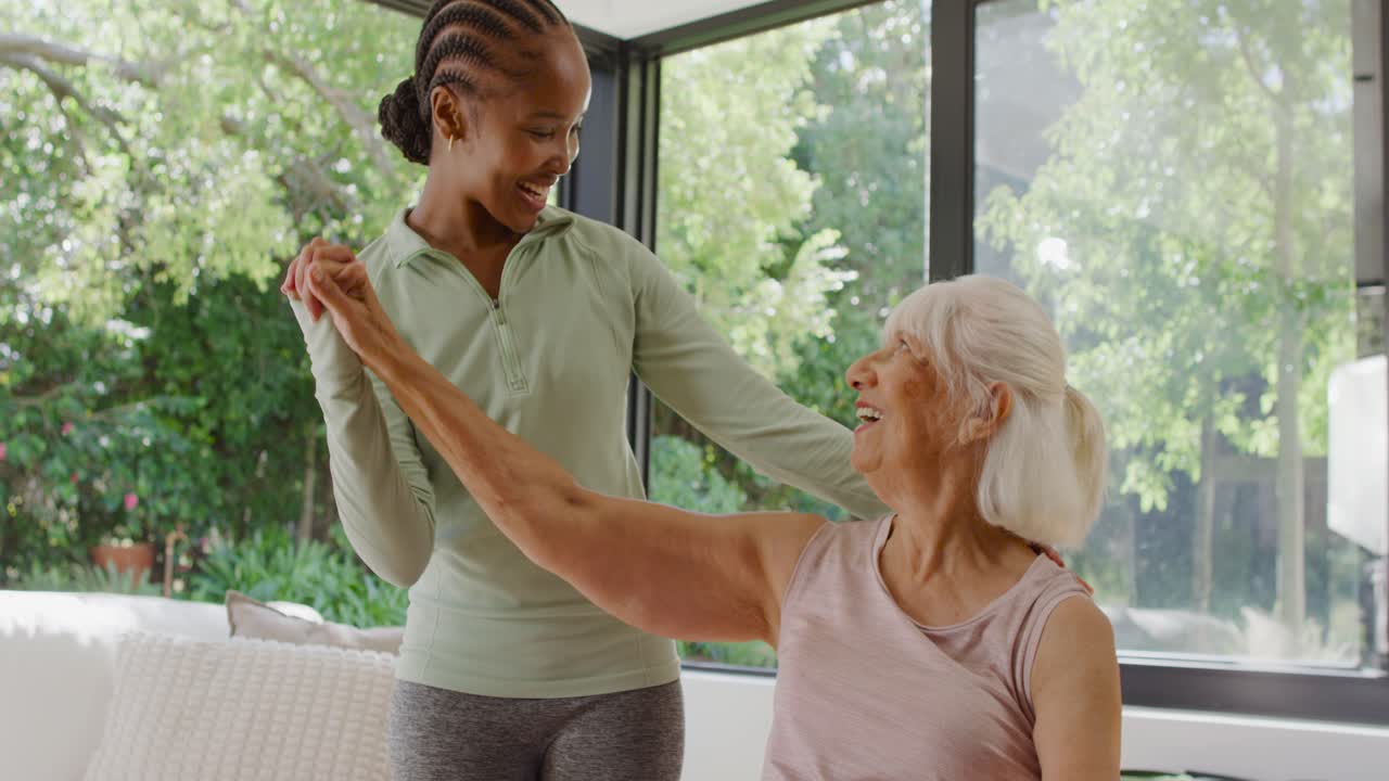 Senior on couch greeting African American aide in green top guiding arm stretch for rehab