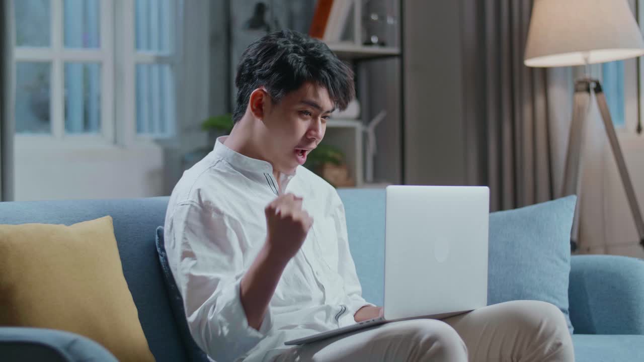 Happy Asian Man Celebrating While Sitting On Sofa And Using Laptop Computer In The Living Room