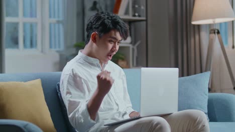 happy asian man celebrating while sitting on sofa and using laptop computer in the living room