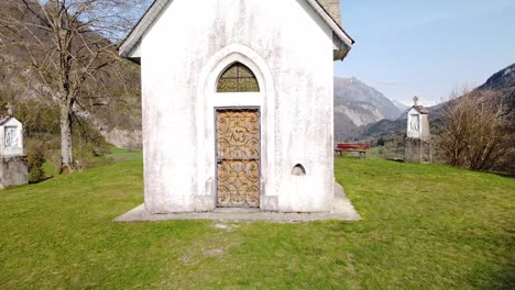 Drone-Shot-of-the-Front-of-a-Chapel-on-top-of-a-Hill-in-front-of-Alpine-Mountains