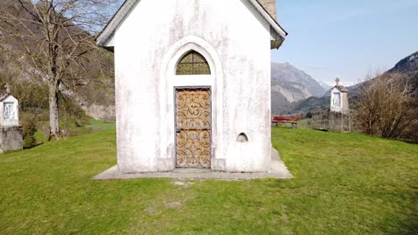 drone shot of the front of a chapel on top of a hill in front of alpine mountains