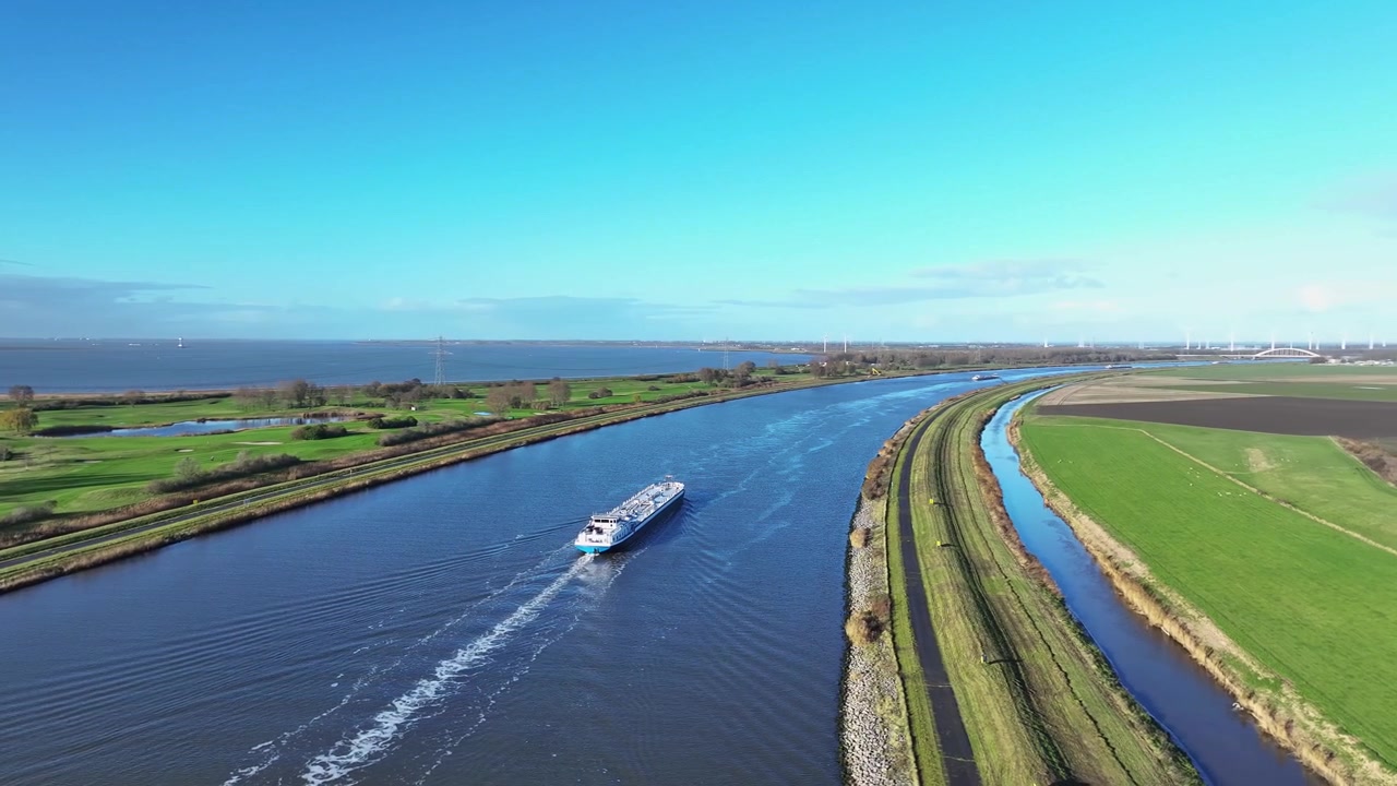 Drohnenaufnahme eines Binnenschiffs, das einen von grasbewachsenen Feldern gesäumten Kanal befährt, mit einem daneben verlaufenden Fluss unter hellem, sonnigem Himmel.