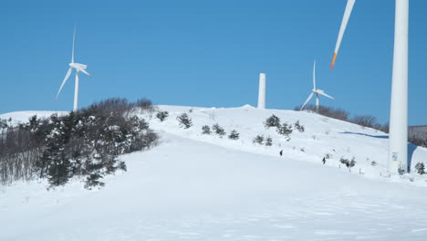 group of hikers on walking trail in winter mountains under gigantic wind turbines at daegwallyeong sky ranch, south korea - wide slow motion landscape