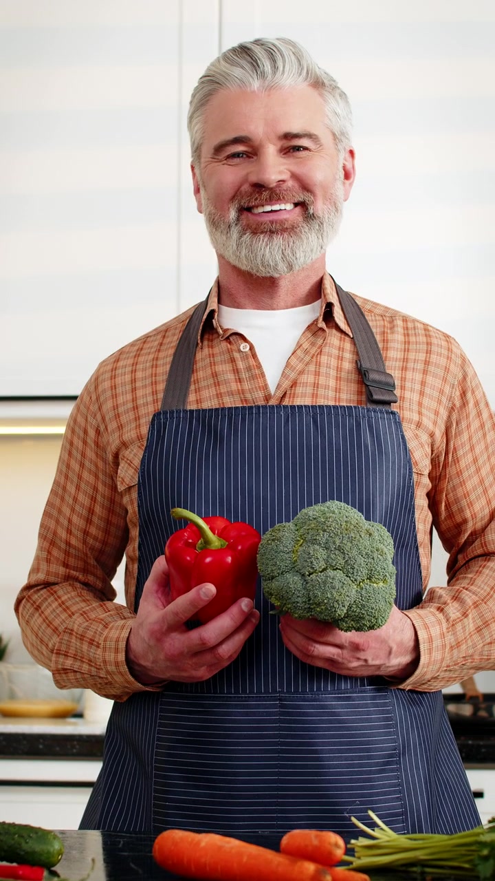 Lachender Mann mittleren Alters hält frische Paprika und Brokkoli zur gesunden Diät-Vorbereitung in der heimischen Küche, fit.