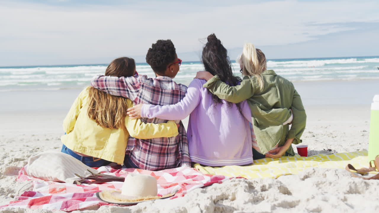 grupo feliz de diversas amigas divirtiéndose, sentadas en mantas, abrazándose en la playa