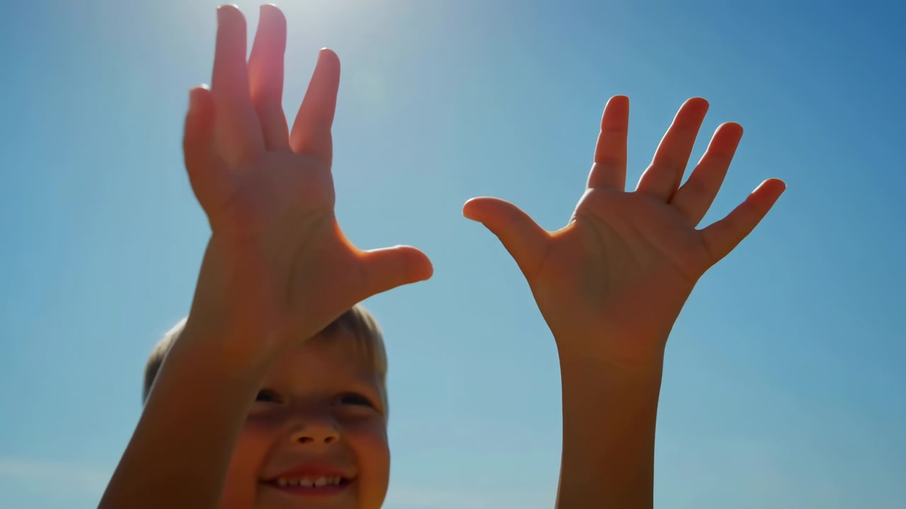 Un bambino sorridente è posizionato in basso a sinistra dell'inquadratura con entrambe le braccia alzate verso il cielo azzurro limpido, illuminato da una forte luce solare con una morbida retroilluminazione