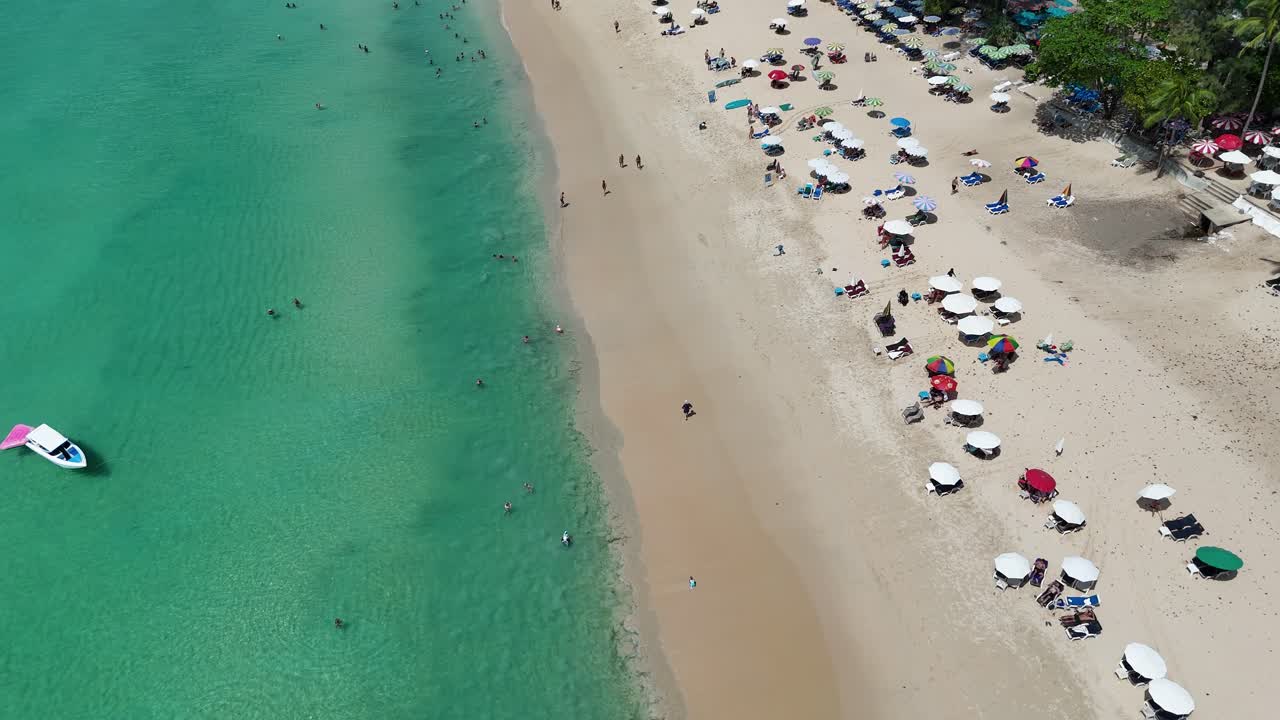 Drone footage captures a vibrant beach in Phuket, Thailand, showcasing turquoise waters, sandy shores, and colorful umbrellas under bright daylight