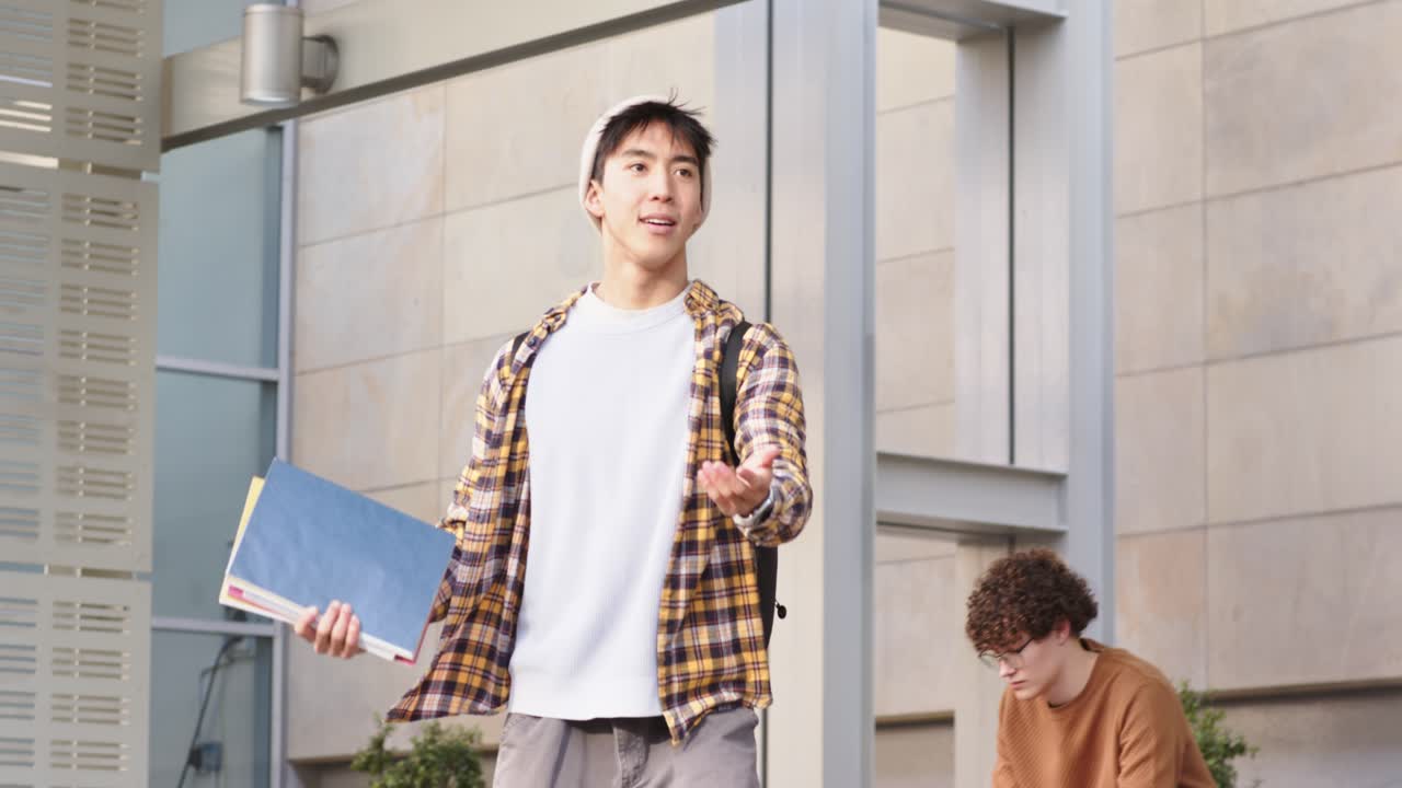 一名亞洲男學生手拿文件夾走過校園庭院，舉手向朋友打招呼。