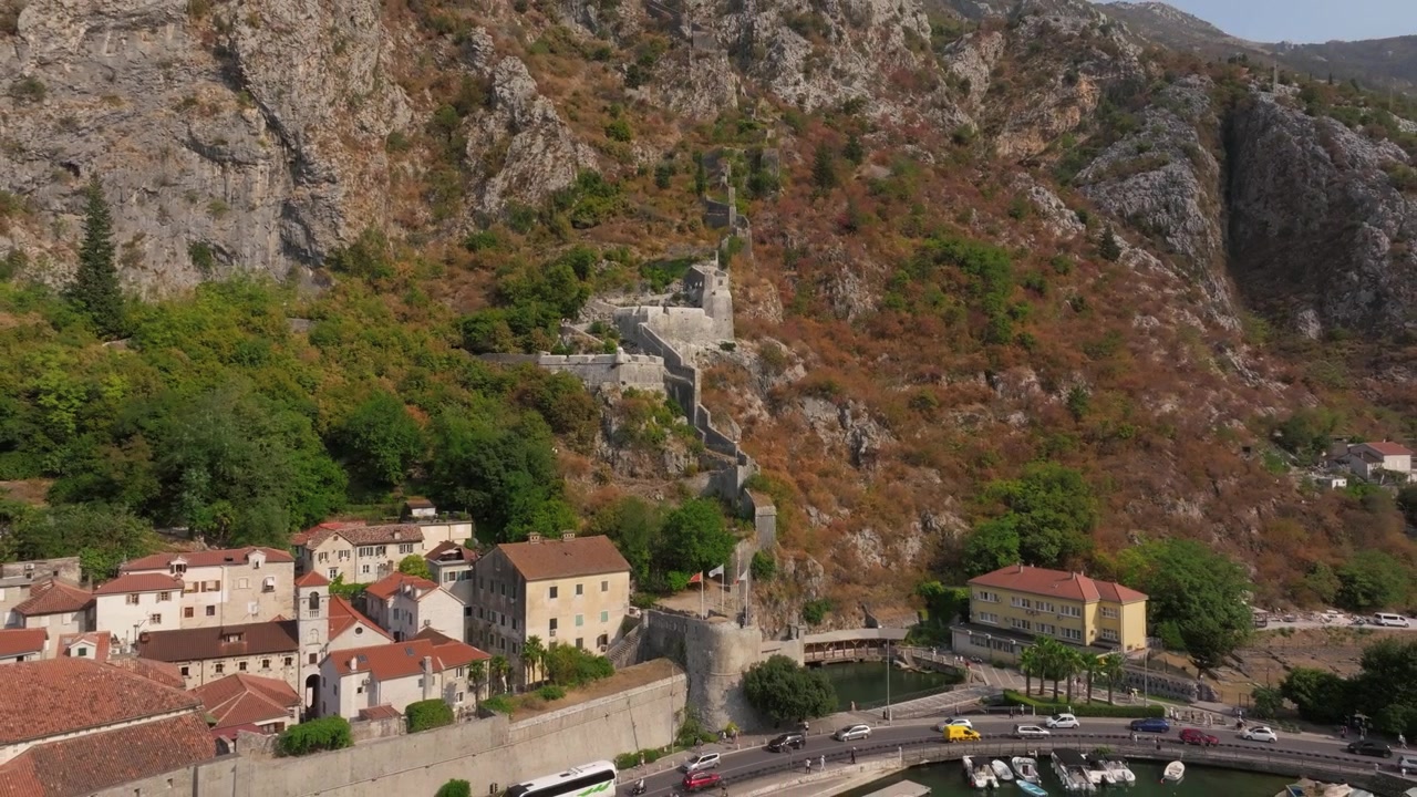 Mura di fortificazione che risalgono un ripido versante montuoso dietro case sul lungomare, traffico e un piccolo bacino portuale in Montenegro
