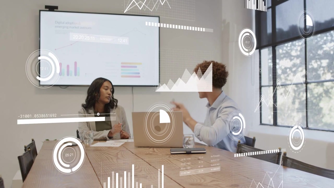 Woman writing, glancing at laptop, peer leaning, pointing, overlays moving on charts for biz review