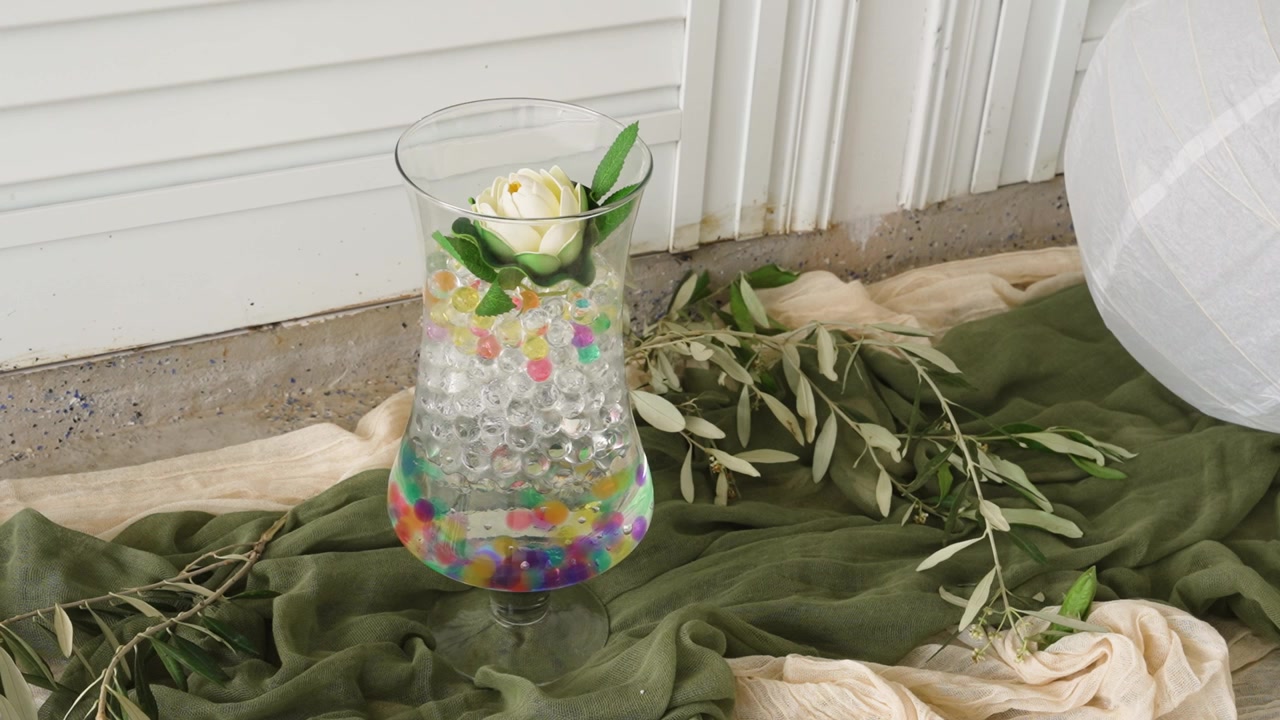 Un bicchiere trasparente contiene una bevanda colorata decorata con perline e un fiore. Il bicchiere poggia su un tessuto morbido con colori verdi e bianco sporco. La vegetazione circonda l'ambiente, completando l'aspetto