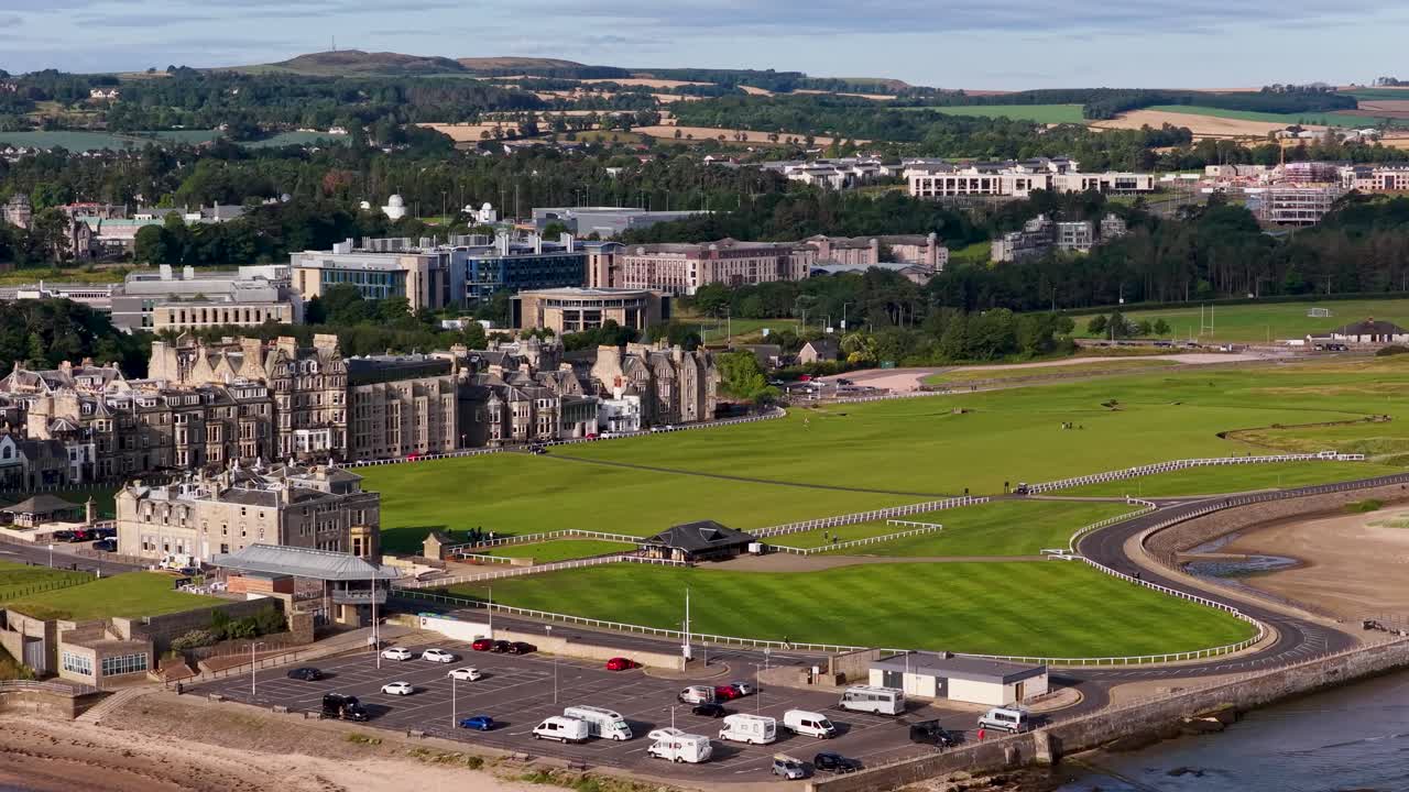 Drone glides over iconic golf course, historic clubhouse, coastal town, and green fairways in daylight