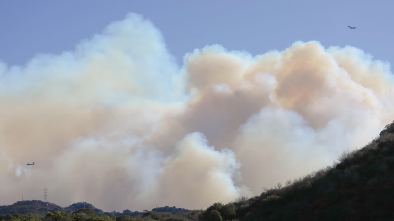 Firefighting planes soaring above dense smoke, blue sky contrasts wildfire destruction below