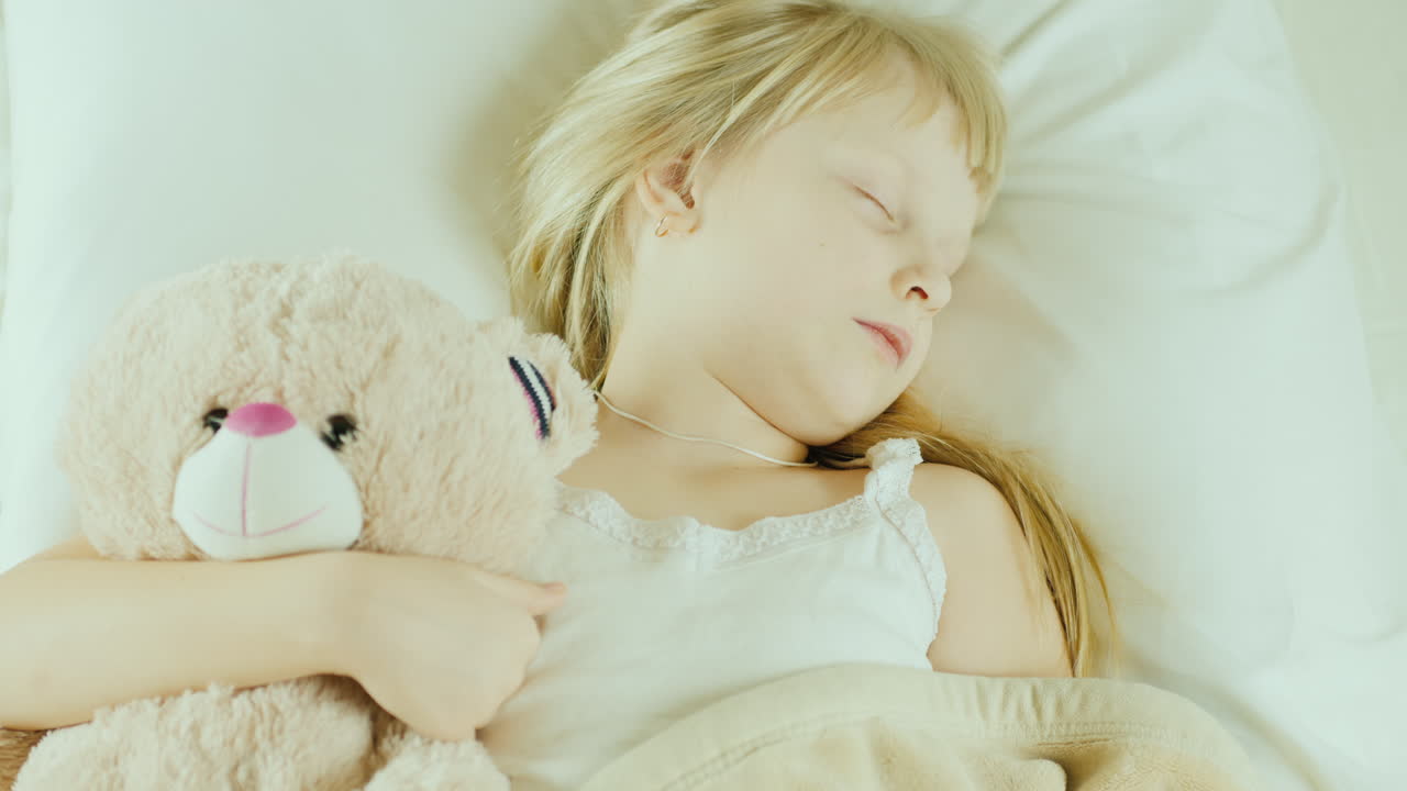 침대에서 자고있는 어린 소녀는 장난감 곰 평면도를 껴안습니다.