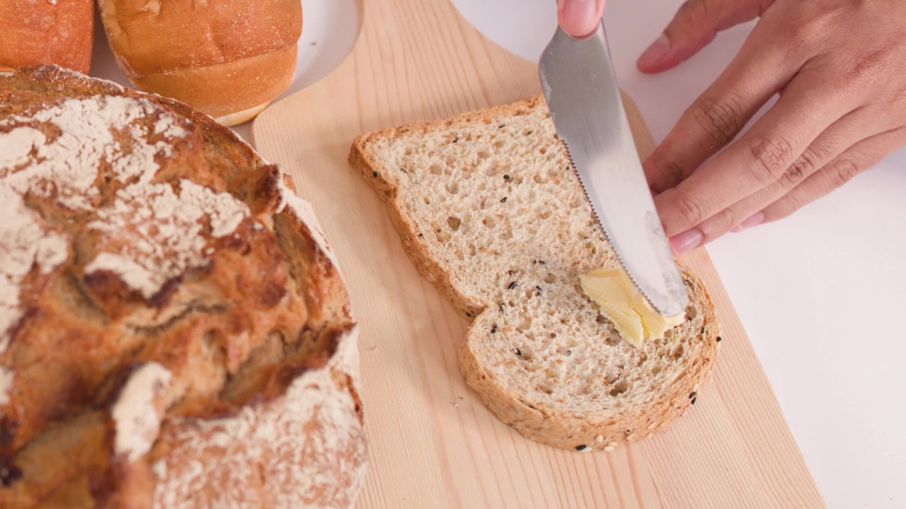 Nærbillede ovenfra af hænder, der smører smør på brød blandt et udvalg af brød