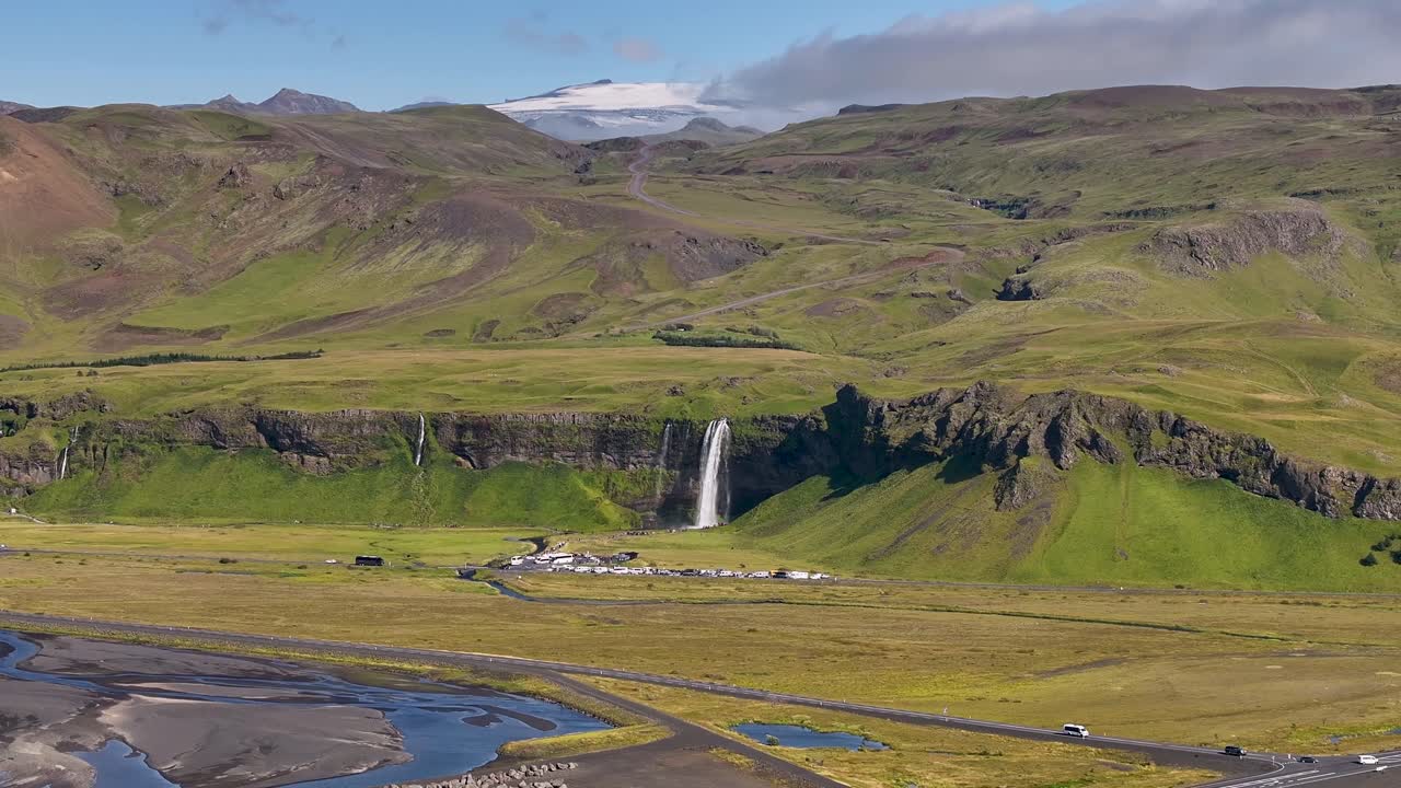 Aerial wide shot of Seljalandsfoss waterfall in idyllic mountain landscape. Sunny day with snowy peaks of hills in Iceland. Panorama view