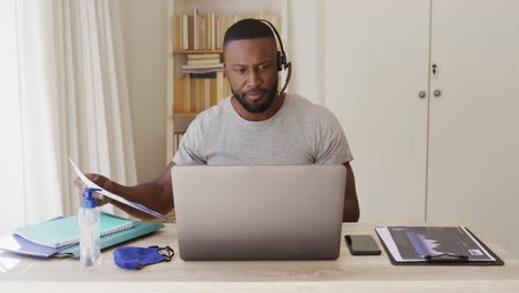 hombre afroamericano en casa con auriculares de teléfono usando una computadora portátil
