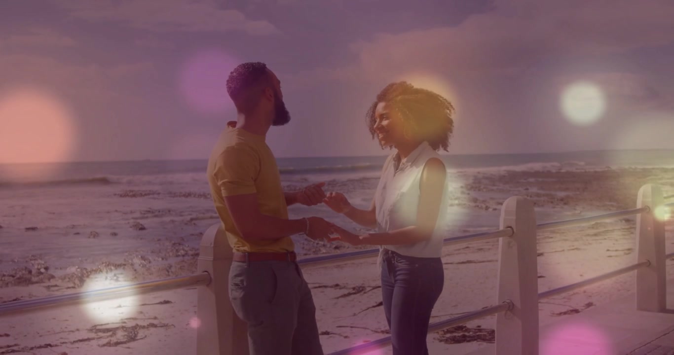 Coppia che si tiene per mano e si avvicina, mentre un effetto bokeh sfocato attraversa la coppia e la ringhiera della spiaggia, per marketing.