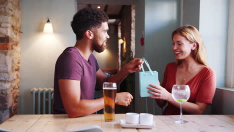 young adult mixed race man giving a present to his girlfriend, sitting with drinks at a table in a pub, close up, side view