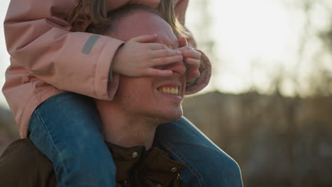 father and daughter playing in the park at sunset, with the daughter sitting on his shoulders, covering his eyes while both laugh joyfully