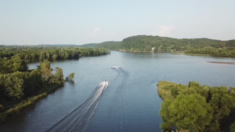 Twin speedboats race past green islands on a wide river, carving white wakes into calm waters surrounded by dense forest and distant hills on a sunny day.