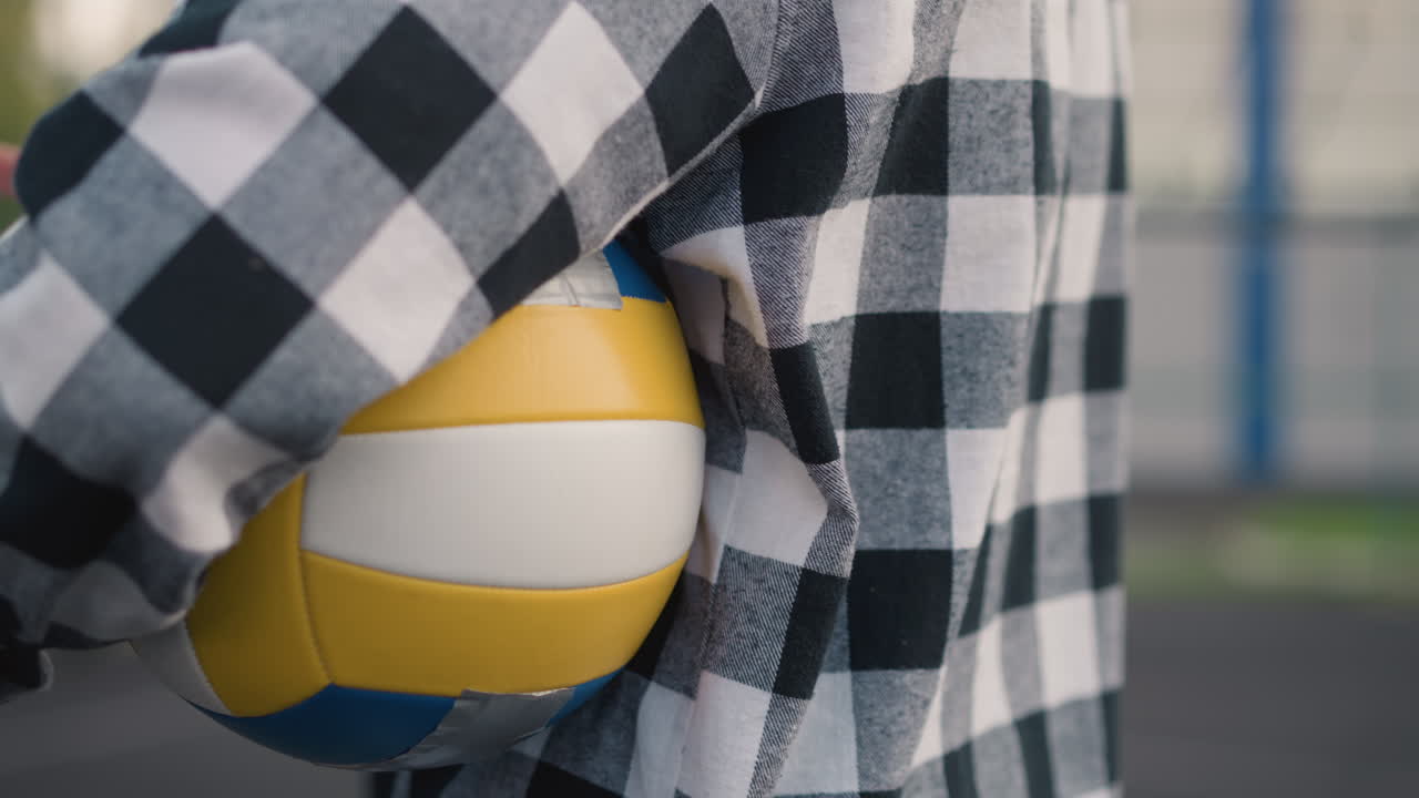 vista lateral de primer plano del entrenador de voleibol con la pelota bajo el brazo y la tarjeta de récord en la mano, con el fondo borroso de la cancha de voleobol al aire libre
