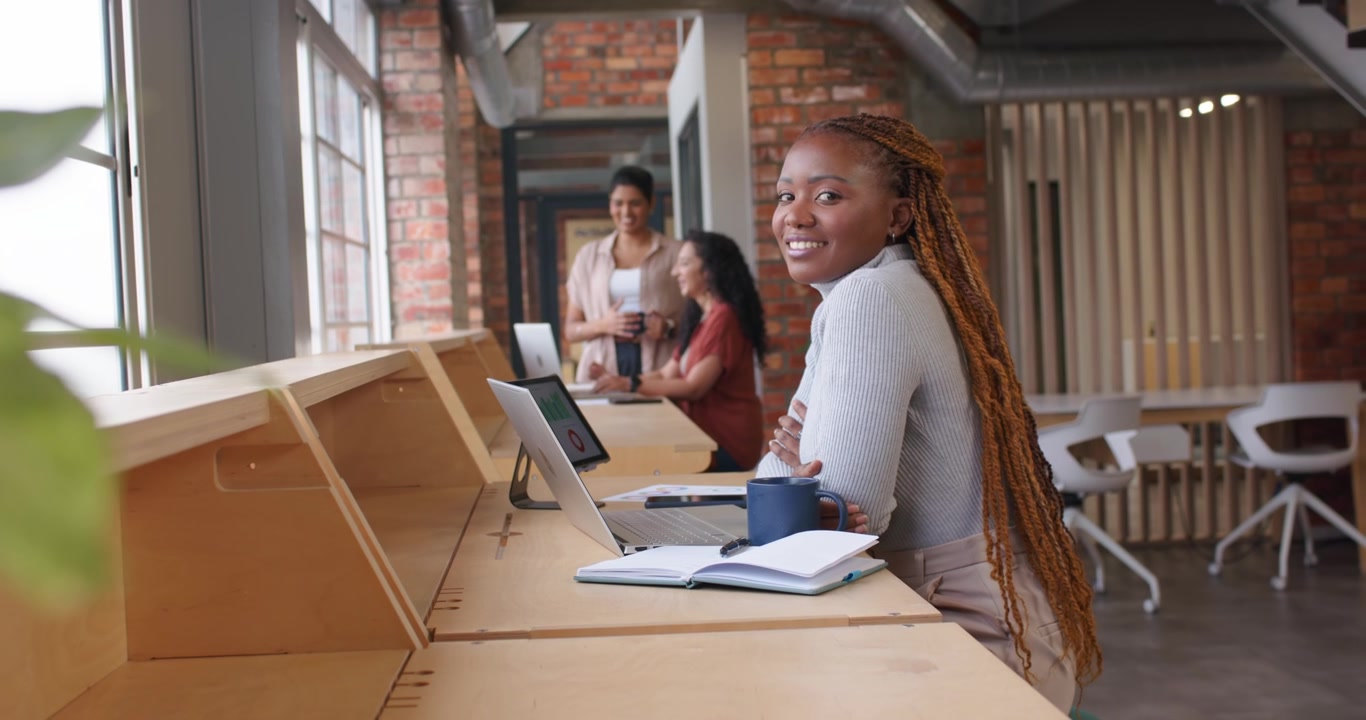 Plant shifting, African-American woman working at counter on laptop, blue mug while peers chatting