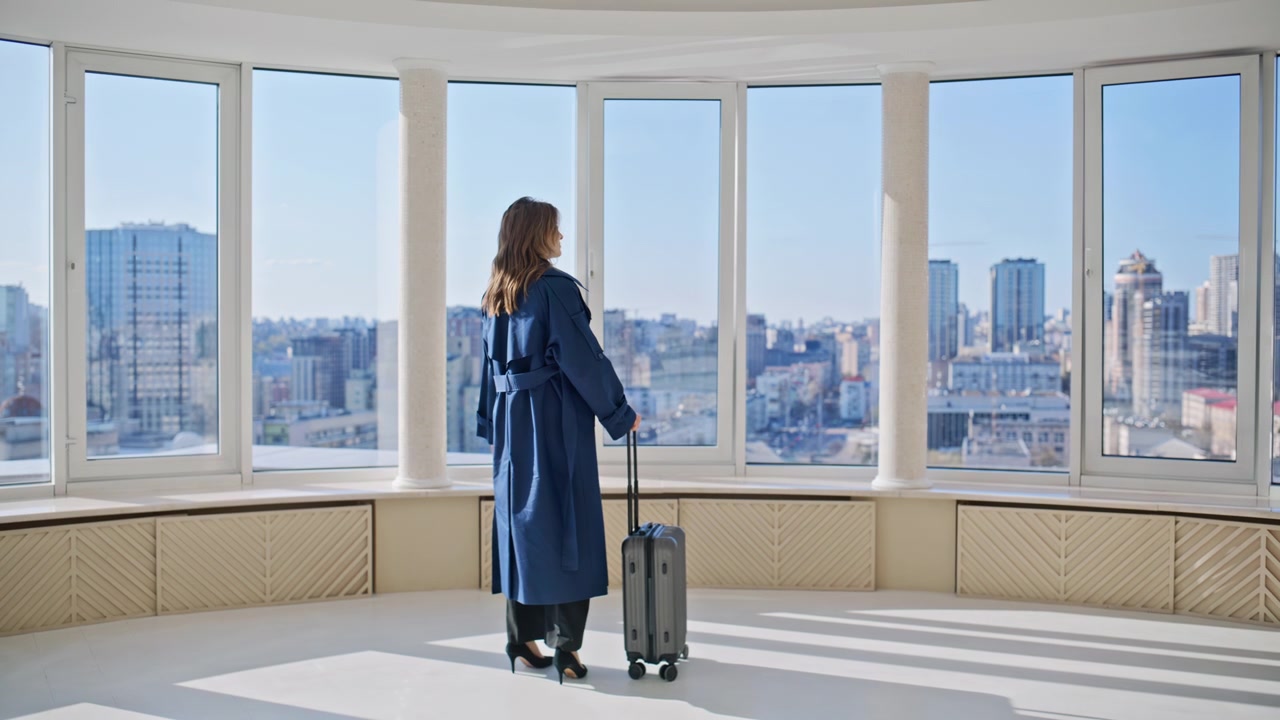 Woman with suitcase empty luxury apartment. Traveler looking city skyline view.