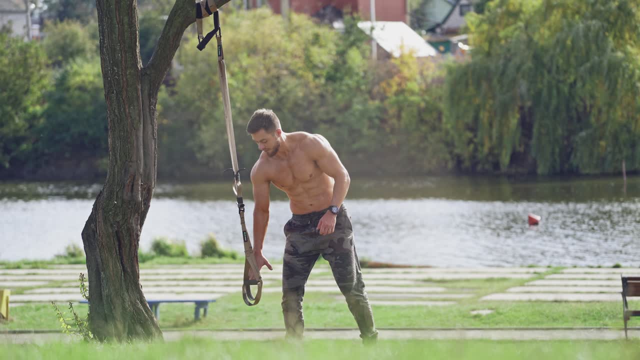 Healthy sportsman begins training on trx outdoors. Active athlete training with straps on the river background in a sunny day.
