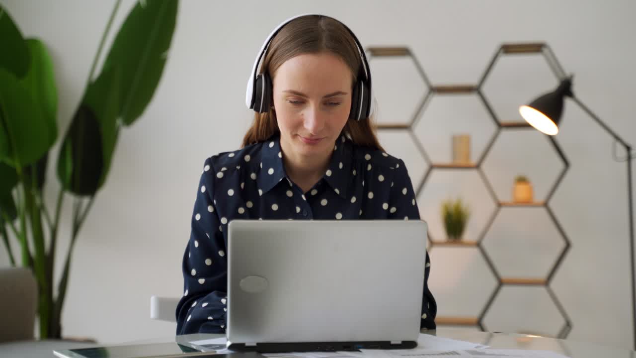 A young female project manager puts on headphones and works on a laptop computer in an office setting