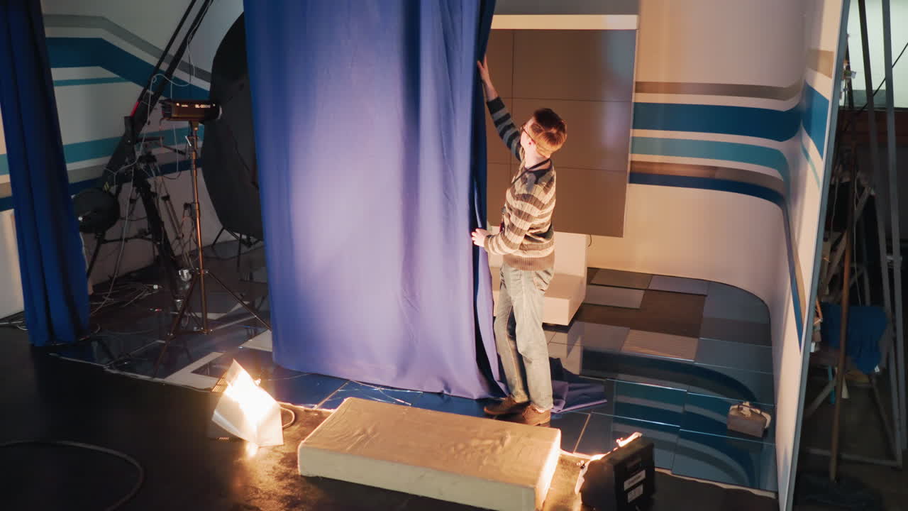Studio technician wearing striped sweater pulls large blue curtain on film set revealing backstage environment with lights, camera tripod, soft,box, wires, platform block, abstract wall graphics